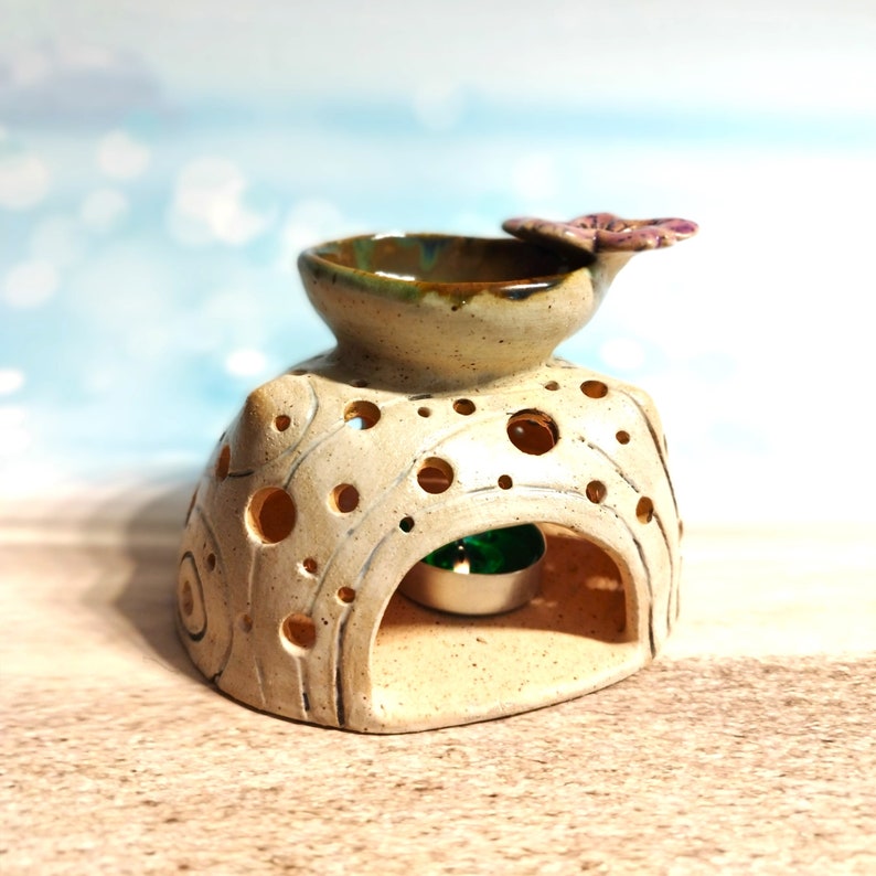 May include: A ceramic oil burner with a white glaze and a decorative butterfly design. The burner has a small bowl on top for essential oils and a hollow space underneath for a tea light.