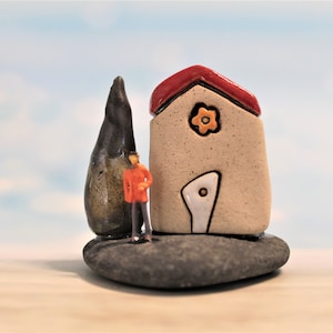 May include: A small ceramic house with a red roof and a white door. The house has a yellow flower on the front. A small figure in a red jacket stands next to the house. The house is sitting on a gray rock.