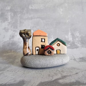 May include: A miniature ceramic village scene featuring three houses, a tree, and a grey stone base. The houses are painted in shades of beige, green, and red. The tree is brown and green.