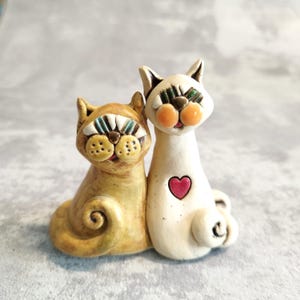 May include: Two ceramic cat figurines, one white with a red heart and the other yellow with blue and green eyes. The cats are sitting side by side.