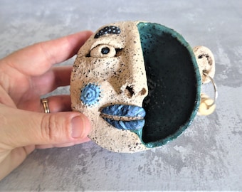 May include: A ceramic wall hanging in the shape of a face with a blue and white painted design. One side of the face is painted teal and has a small hole for hanging.