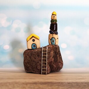 May include: A miniature ceramic house with a yellow roof and blue door sits on a brown rock next to a taller ceramic tower with a yellow top and blue accents. A small silver ladder is leaning against the rock.