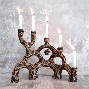 May include: A black and brown ceramic candle holder with a tree branch design. The holder has five candle holders and is decorated with leaves and a small flower.