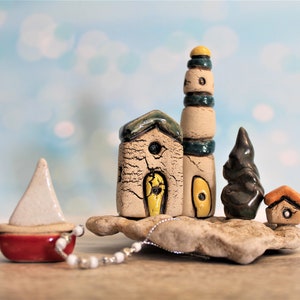 Puede incluir: Una escena de pueblo en miniatura de cerámica con un faro, casas y un pequeño velero. Las casas están pintadas en tonos de blanco, amarillo y marrón, con acentos verdes y azules. El faro es alto y delgado, con una parte superior amarilla y rayas azules y verdes. El velero es rojo con una vela blanca. La escena está colocada sobre una base rocosa.