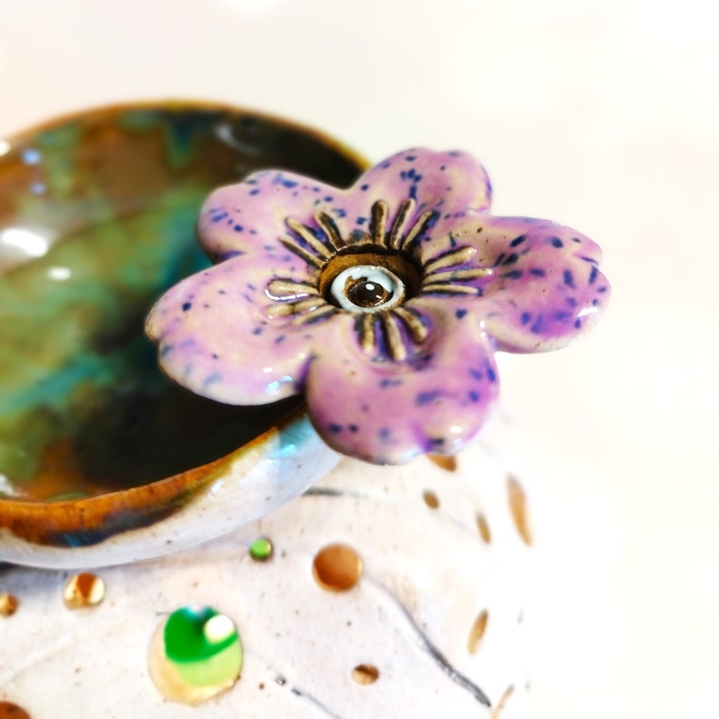May include: A ceramic diffuser with a purple flower on top. The diffuser has a white glaze with brown and green accents. The flower has a brown centre with a white dot in the middle.