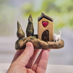 May include: A small ceramic sculpture featuring a tan house with a dark brown arched door and a red heart cutout above it. Two green, textured, cone-shaped trees stand to the left of the house, and a small white cat figurine sits on the right.