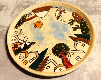 May include: A round ceramic plate with a whimsical painted design of a town with houses, trees, a windmill, and a sun. The plate has a beige background with brown, green, blue, and yellow accents.