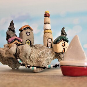 May include: A ceramic miniature village scene with a lighthouse, houses, and a sailboat. The houses are painted in various colors, including brown, beige, and green. The sailboat is white with a red hull. The scene is set on a rocky base.