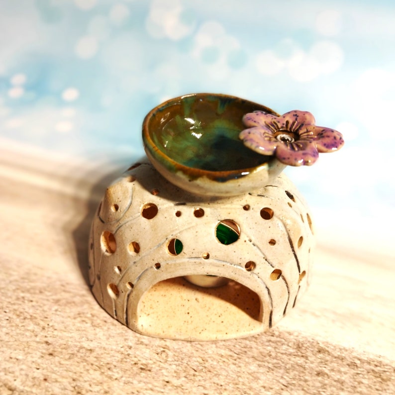 May include: A ceramic oil burner with a white base and a green and brown bowl on top. The bowl has a purple flower on the side. The base has a hole in the centre for a tea light.