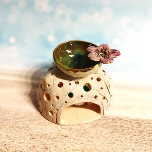 May include: A white ceramic oil burner with a green and brown bowl on top and a purple flower accent. The burner has a decorative design with holes and a small opening at the front.