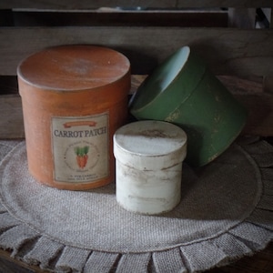 May include: Three round cardboard boxes, one orange with the text "Carrot Patch" and a picture of carrots, one green, and one white, on a brown burlap surface with a ruffled edge.