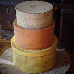 Può includere: Tre scatole rotonde di legno impilate l'una sull'altra. La scatola superiore è bianca, la scatola centrale è arancione e la scatola inferiore è gialla. Le scatole sono su un tappeto marrone intrecciato.