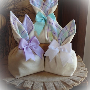 Peut inclure: Trois sacs cadeaux de lapin de Pâques en toile de jute avec des oreilles de lapin à carreaux. Les sacs sont attachés avec des nœuds en lavande, bleu clair et blanc.