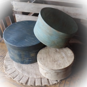 May include: Three round wooden boxes stacked on top of each other. The top box is a light blue-green color, the middle box is a dark blue color, and the bottom box is a light beige color. The boxes are sitting on a brown burlap rug with a ruffled edge.