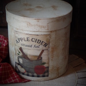 Op de afbeelding: Een witte cilindrische container met een verweerde afwerking en een label met de tekst "Apple Cider served hot". Het label toont een vintage illustratie van een mok appelcider met kaneelstokjes en een rode appel.