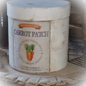 Könnte beinhalten: Ein weißer zylindrischer Behälter mit einem Vintage-Etikett, auf dem "McGregor Farms Carrot Patch" steht. Das Etikett zeigt eine handgezeichnete Illustration von Karotten und den Text "Hand Picked Fresh Daily Locally Grown Organic 5¢ Per Carrot One Mile Ahead".