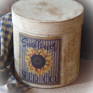 May include: A white cylindrical container with a distressed finish and a label that reads "Sunflower Seed Co." The label features a sunflower with yellow petals and a dark brown center. The container is sitting on a brown burlap surface.