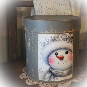 May include: A round, distressed grey storage box featuring a smiling snowman illustration. The snowman has a carrot nose, rosy cheeks, and a white fur-trimmed hat and scarf. The box rests on a cream lace doily.