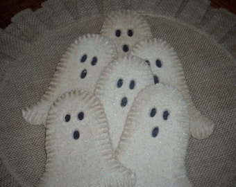 May include: Five white ghost-shaped fabric ornaments with black eyes and mouths, arranged on a brown burlap surface.
