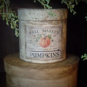 May include: A white round tin container with a distressed finish and a label that reads "Fall Market" with a picture of pumpkins and the words "You Pick" and "Pumpkins".