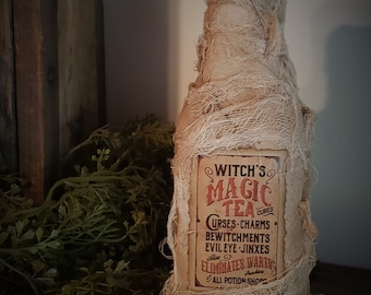Può includere: Una bottiglia di vetro marrone avvolta in juta con un'etichetta che recita "Witch's Magic Tea Cures Curses, Charms, Bewitchments, Evil Eye, Jinxes, Also Eliminates Warts, Purchase All Potion Shops".