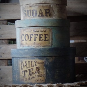 Könnte beinhalten: Drei vintage, runde, bemalte Blechdosen mit Deckeln. Die oberste Dose ist weiß mit schwarzer Beschriftung, die "SUGAR" lautet. Die mittlere Dose ist blau mit weißer Beschriftung, die "Finest Roasted COFFEE" lautet. Die unterste Dose ist dunkelblau mit einem braunen Etikett, auf dem "DAILY Brand TEA Finest Orange Pekoe" steht.