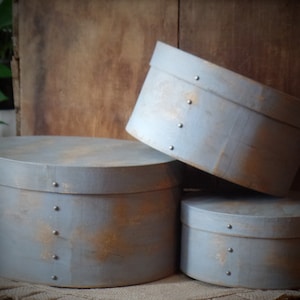 May include: Three round, gray painted wooden boxes with metal accents. The boxes are stacked on top of each other, with the largest box on the bottom and the smallest box on top.