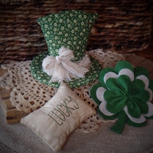 以下が含まれることがあります： 緑と白の布製の帽子に白いレースのリボンが飾られ、小さな茶色の麻の枕には「Lucky」という刺繍が施されています。帽子と枕は、クロシェ編みのドイリーの上に置かれています。