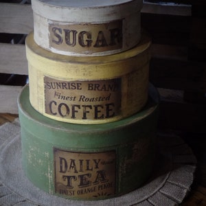 May include: Three vintage-style round canisters with lids, stacked on a hessian surface. The top canister is white with the word "SUGAR" in brown lettering. The middle canister is yellow with the words "SUNRISE BRAND Finest Roasted COFFEE" in brown lettering. The bottom canister is green with the words "DAILY TEA Finest Orange Pekoe" in brown lettering.