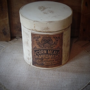 May include: A white, round, vintage-style container with a brown label that reads "Corn Meal, Since 1848, Appleton Mills, Finest Yellow Corn, Satisfaction Guaranteed".