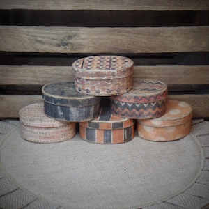 May include: Six round vintage boxes with various patterns and designs. The boxes are stacked in a pyramid shape on a brown burlap rug with a ruffled edge. The boxes are made of wood and have a distressed look.