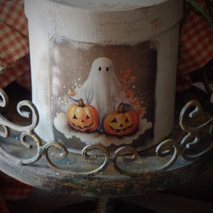 May include: A white round box with a vintage Halloween design featuring a ghost and two jack-o'-lanterns. The box is sitting on a decorative metal stand.