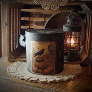 May include: A black, cylindrical storage container with a painted scene of a crow perched on pumpkins. A lit lantern and wooden crate are in the background. The container sits on a burlap placemat, creating a rustic, autumnal theme.
