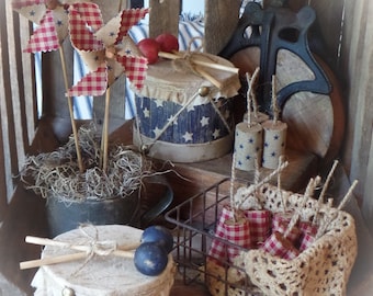May include: A patriotic themed display with two drums, one decorated with red, white, and blue stripes and the other with stars and stripes. There are also pinwheels and firecrackers in red, white, and blue.