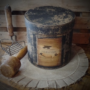 Peut inclure: Une boîte ronde en métal noir et brun avec une étiquette indiquant "Dealer in Fresh & Salt Meats". La boîte est posée sur un dessous de plat en toile de jute à bord festonné.