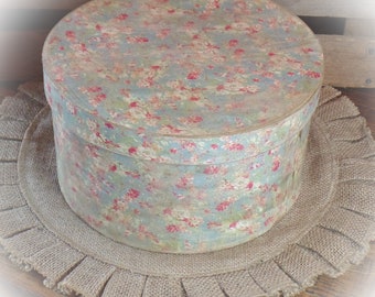 May include: A round, vintage hatbox with a floral pattern in shades of blue, pink, and white. The hatbox is sitting on a brown burlap mat with a ruffled edge.