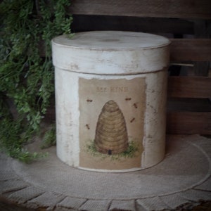 May include: White cylindrical container with a distressed finish and a printed image of a beehive with the text "Bee Kind".