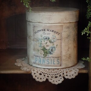 May include: A white cylindrical container with a vintage label that reads "Flower Market Est. 1954" and "Pansies" with a floral design featuring blue and white pansies.