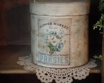 Puede incluir: Un recipiente cilíndrico blanco con una etiqueta vintage que dice "Flower Market Est. 1954" y "Pansies" con un diseño floral con pensamientos azules y blancos.