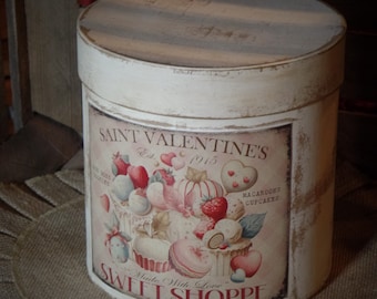 Puede incluir: Caja cilíndrica de hojalata blanca con un diseño vintage. La caja presenta una ilustración roja y blanca de un pastel con fresas, macarons y cupcakes. El texto en la caja dice "Saint Valentine's Est. 1915 Swift Shoppe Made With Love".