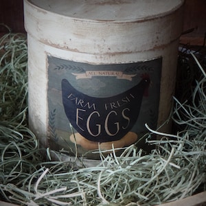 Może przedstawiać: Biały, zużyty, cylindryczny pojemnik z etykietą z napisem "All Natural Farm Fresh Eggs". Pojemnik stoi na zielonym słomie.