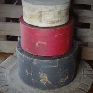 Puede incluir: Tres cajas de madera redondas apiladas una encima de la otra. La caja superior es blanca, la caja del medio es roja y la caja inferior es gris oscuro. Las cajas están todas desgastadas y tienen un aspecto rústico. Las cajas están sobre una alfombra de arpillera.