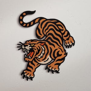 May include: A wooden tiger cutout with orange and black stripes, a white belly, and an open mouth. The tiger is in a crouching position, with its claws extended. The artwork is against a white background.