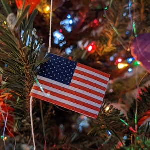 May include: A small American flag with red and white stripes and a blue field of white stars is attached to a Christmas tree. The tree is decorated with colorful lights and ornaments, creating a festive holiday scene.