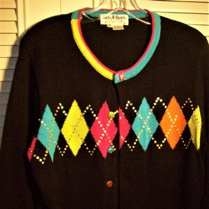 May include: Black cardigan sweater with a colorful argyle pattern. The sweater features turquoise, yellow, orange, and pink diamond shapes, embellished with sequins. The trim is turquoise, pink, and yellow. The buttons are multi-colored.