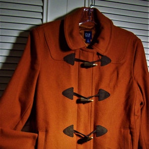 May include: A rust-colored wool coat with a brown collar and four brown toggle closures. The coat has a label that says "GAP" on the front.