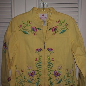 May include: A yellow jacket with a stand-up collar and a full-length zipper. The jacket is adorned with embroidered floral and butterfly designs in shades of pink, purple, green, and blue. The jacket is hanging on a clear plastic hanger.