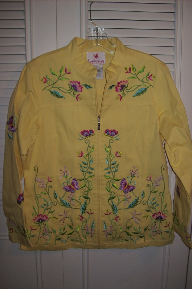 May include: A yellow jacket with a stand-up collar and a full-length zipper. The jacket is adorned with embroidered floral and butterfly designs in shades of pink, purple, green, and blue. The jacket is hanging on a clear plastic hanger.