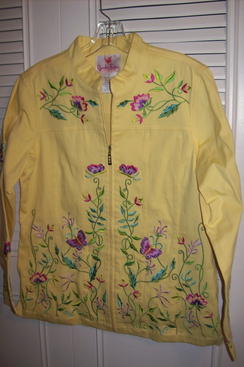 May include: A yellow jacket with a full-length zipper and floral embroidery. The embroidery features purple flowers, green leaves, and butterflies. The jacket is hanging on a clear plastic hanger.