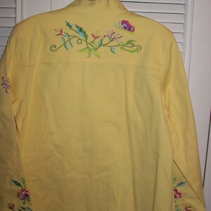 May include: A yellow jacket with long sleeves and a collar. The back of the jacket features embroidered floral and butterfly designs in pink, green, and blue. Additional floral embroidery is on the sleeves.